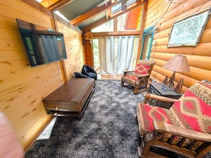 A log cabin interior with a flat-screen TV mounted on the wall, a wooden bench, two patterned armchairs, and a small table with a lamp.