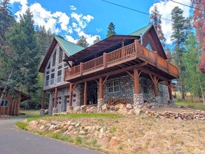 A two-story stone cabin with a wooden balcony, surrounded by a stone wall and a forested landscape.