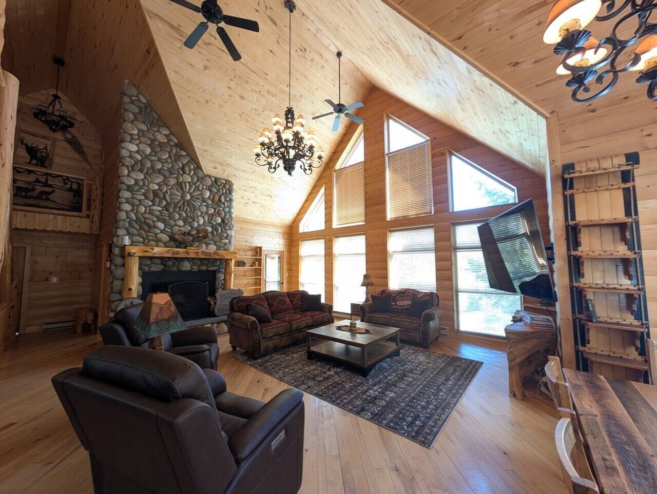 A spacious log cabin interior with a stone fireplace, multiple seating areas, and large windows.