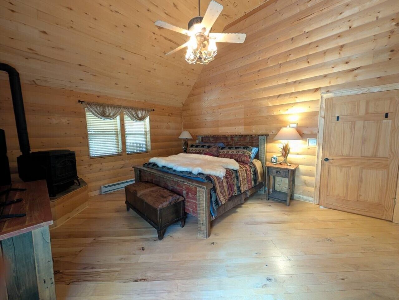 Une chambre dans un chalet en rondins, avec un lit, un radiateur, une fenêtre avec des rideaux, un ventilateur de plafond et une porte.