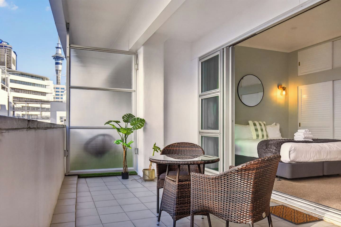 A balcony with a view of a cityscape, a bed with a white headboard, and wicker furniture.