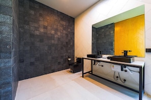 Une salle de bain moderne avec des murs recouverts de carreaux foncés, un plan de toilette blanc et un grand miroir agrémenté d’une touche de jaune.