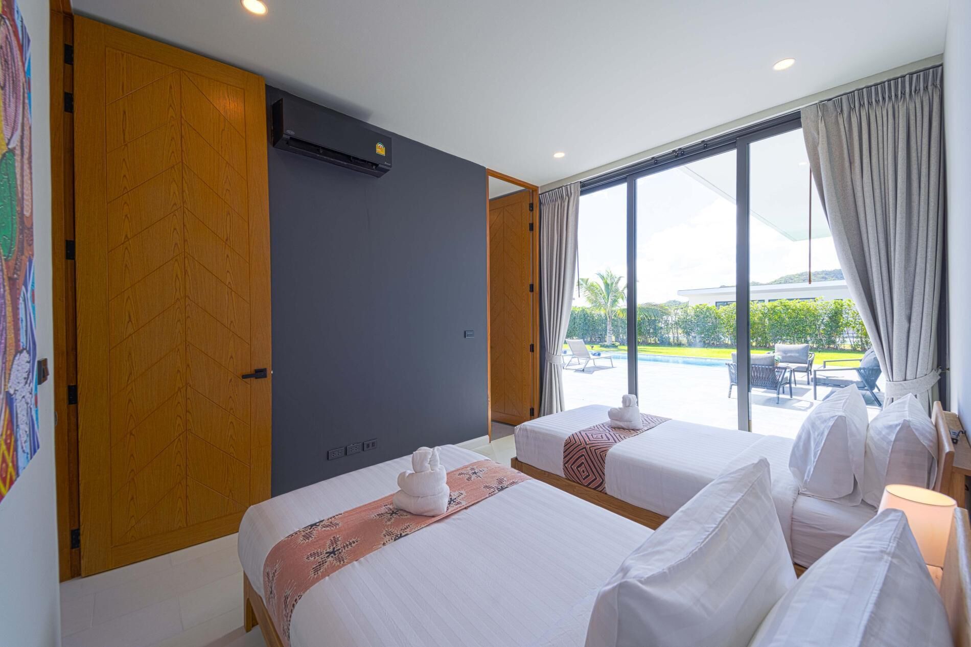 A hotel room with two beds, a large window with curtains, an air conditioning unit, and a view of a swimming pool and greenery outside.