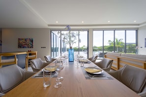Un espace de repas moderne, avec une longue table en bois dressée pour un repas, entourée de fauteuils en cuir. De grandes fenêtres offrent une vue sur l’extérieur.
