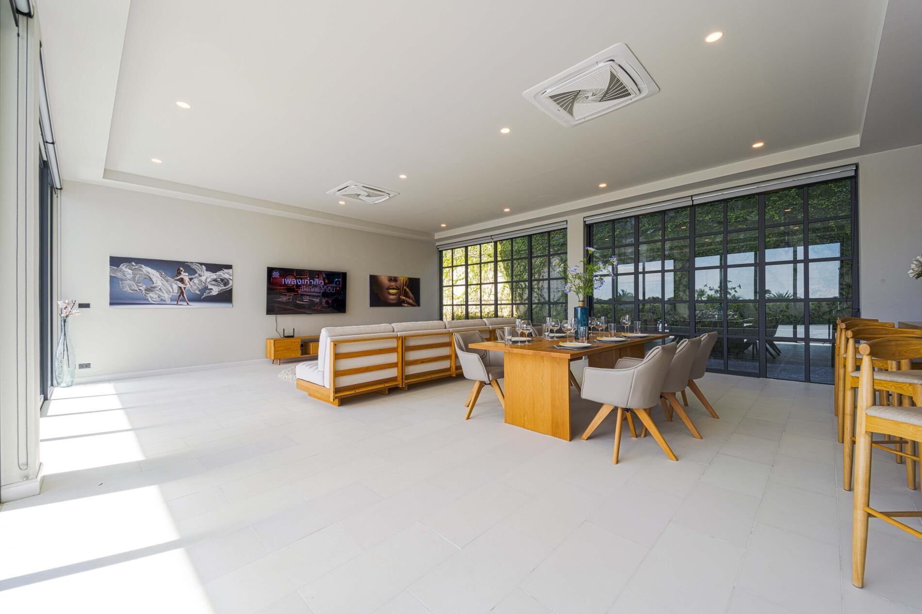 A spacious living area with a sofa, dining table, and chairs, large windows with a view of greenery, and artwork on the walls.