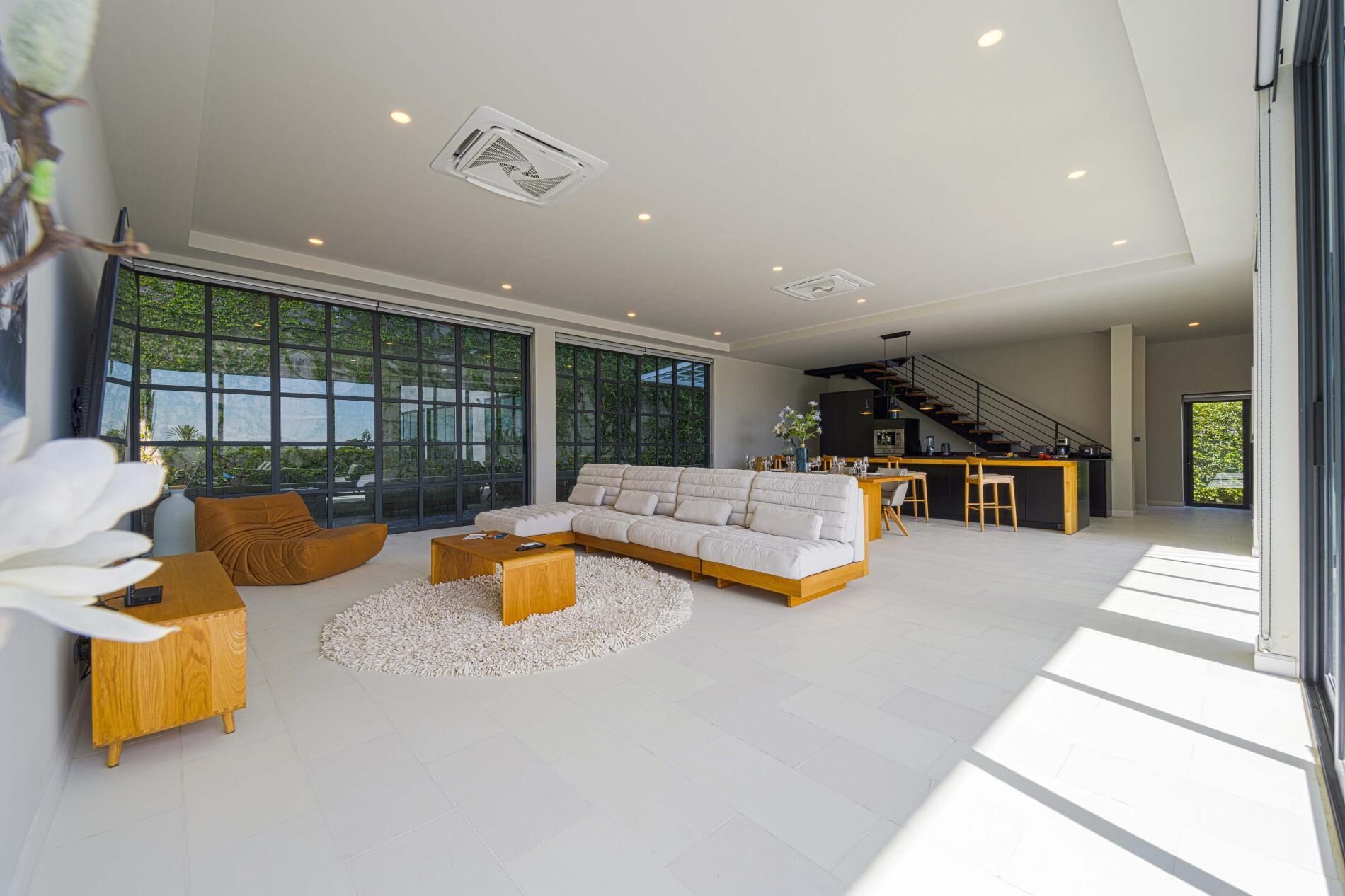 A spacious living room with a large sectional sofa, a wooden coffee table, and a round rug. There is a kitchen area with a bar and a staircase leading to an upper level.