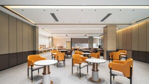 Un hall d’hôtel moderne avec des tables rondes et des chaises orange, un espace de réception doté d’un comptoir, et une grande fenêtre laissant entrer la lumière naturelle.