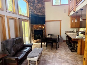A cozy living room with a fireplace, a leather sofa, a dining table, and a kitchen area with wooden cabinets and bar stools. - Luxury 5BD- 4BA in charming Lakewoods Village near Lake of the Woods Crater Lake (Klamath Falls)