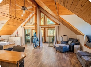 Un'ampia stanza in stile loft con soffitto alto, travi a vista, zona pranzo, angolo salotto con divano e poggiapiedi, e un'ampia porta scorrevole in vetro che si apre su uno spazio esterno.