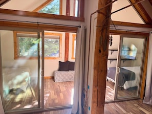 A room with a wooden ceiling, a bed with a white mattress, a window with a view of trees, and a sliding door leading to another room.