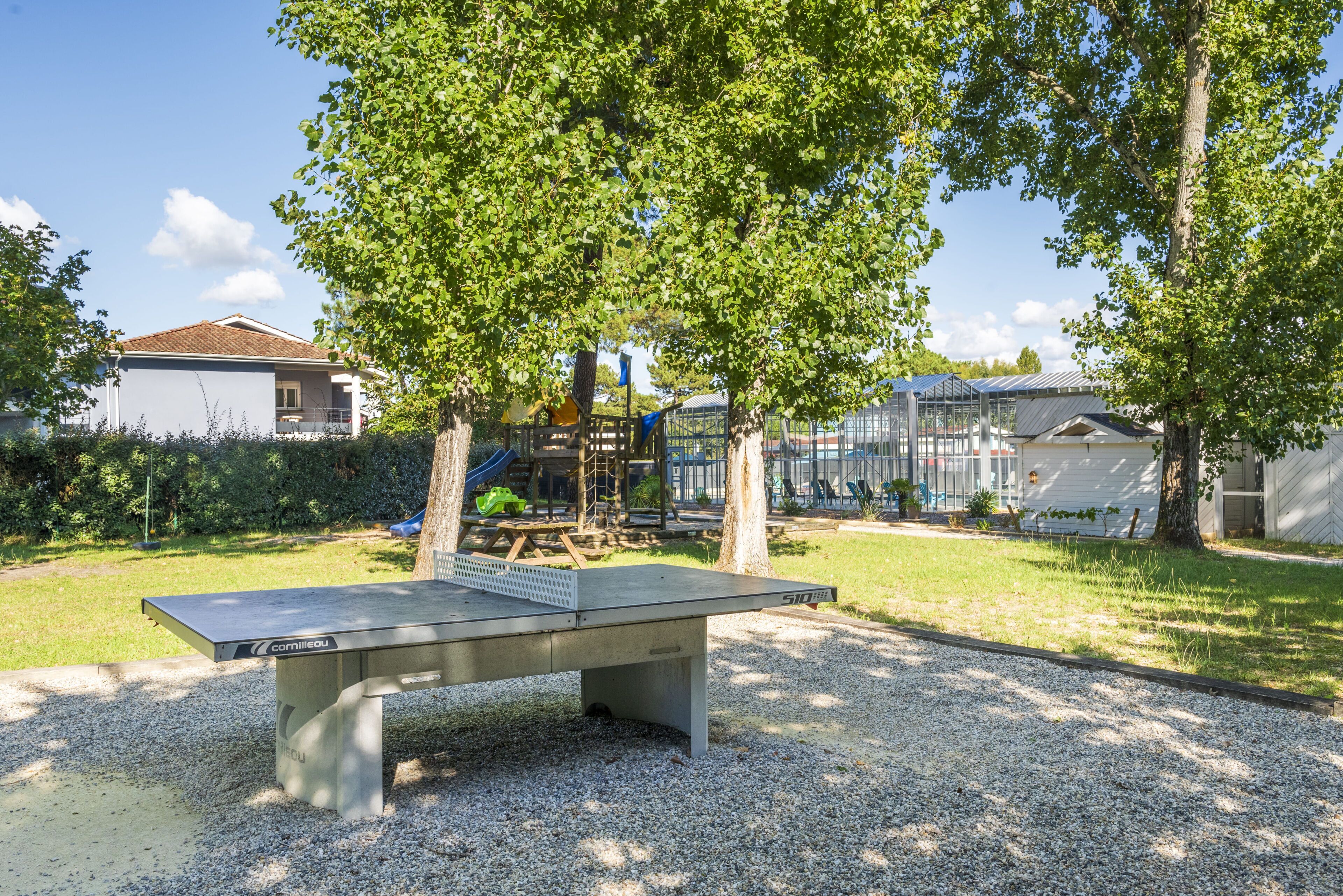 A ping pong table in a park with trees and a playground in the background.