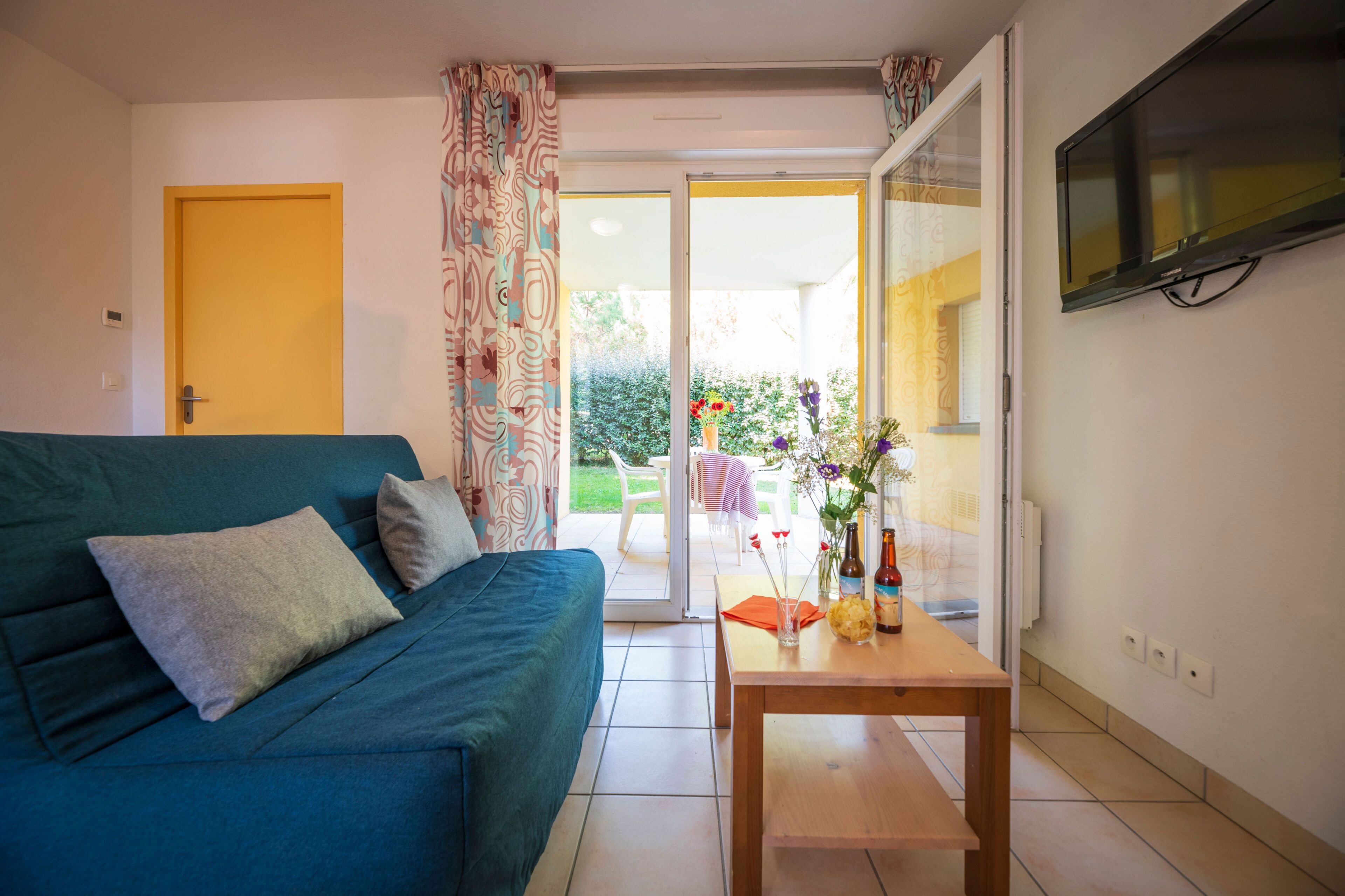 Una sala de estar con un sofá, una mesa con un florero y botellas, y un televisor montado en la pared.