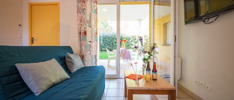 Una sala de estar con un sofá, una mesa con un florero y botellas, y un televisor montado en la pared.