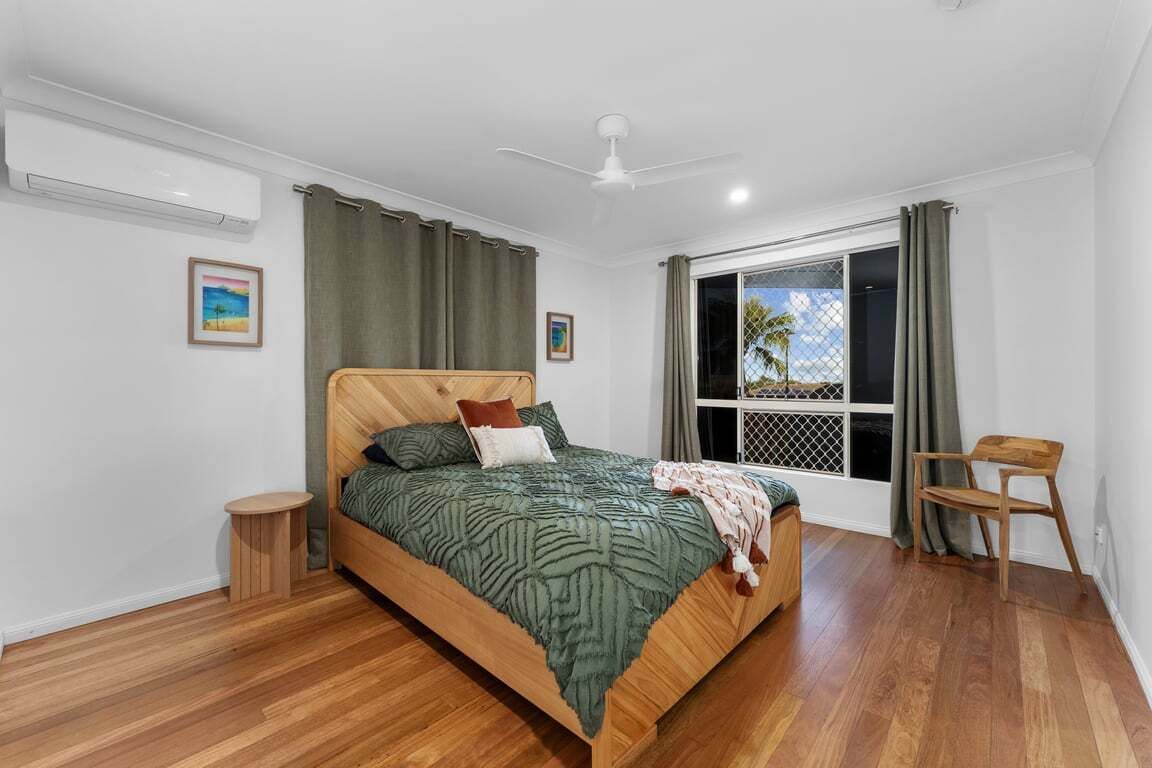 A bedroom with a wooden bed, a small round table, a chair, a ceiling fan, and a window with curtains.