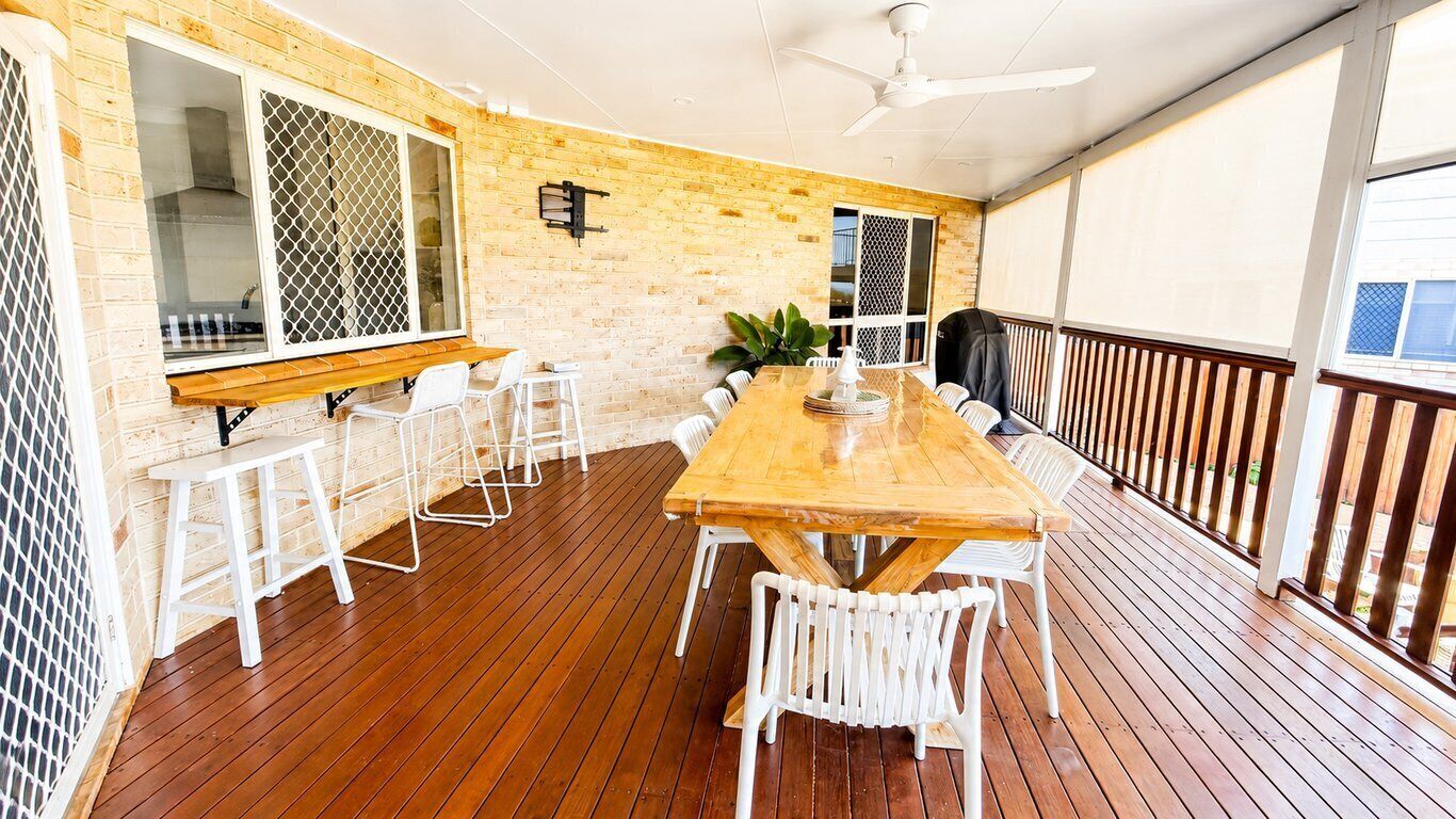 A covered outdoor dining area with a wooden table and white chairs, a ceiling fan, and a brick wall.