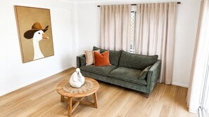 Een woonkamer met een groene bank, een ronde houten salontafel en een schilderij van een eend met een cowboyhoed.