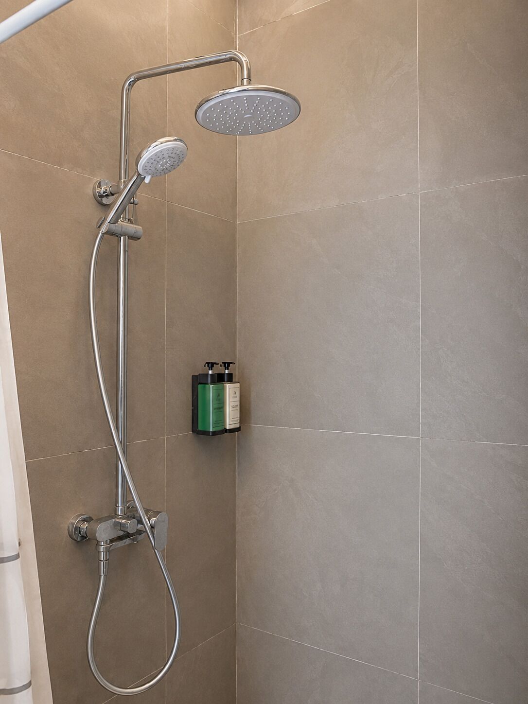 A shower with a handheld showerhead and a fixed overhead showerhead, a handheld shower hose, and a soap dispenser on the wall.