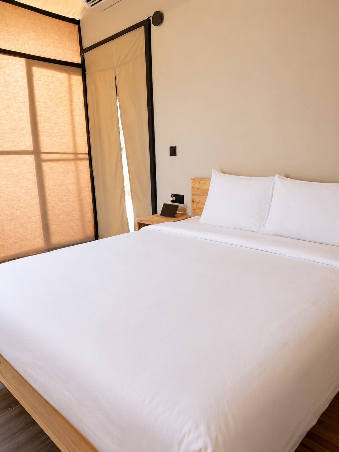 Een net opgemaakt bed met witte lakens, een houten hoofdbord en een klein nachtkastje met een lamp en een stopcontact.