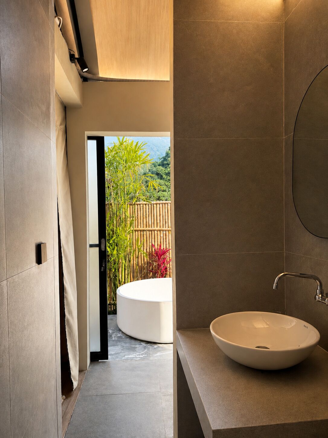 A modern bathroom with a large sink, a mirror, and a view of a bamboo fence and greenery outside.