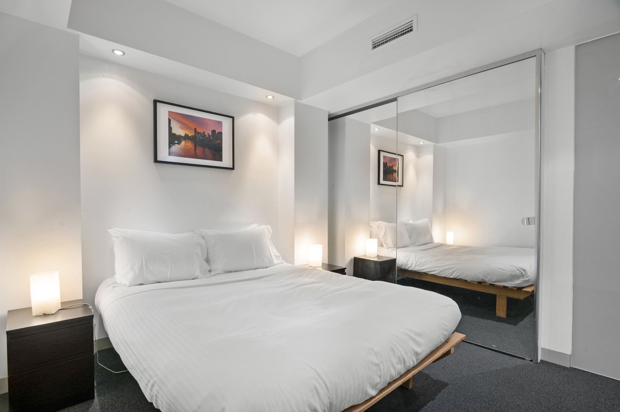 A modern hotel room with a large bed, bedside lamps, a cityscape painting, and a mirror reflecting the room.