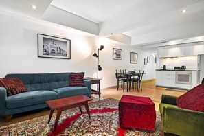 A modern living room with a sofa, coffee table, and ottoman, a dining area with chairs, and a kitchen with appliances. - Melbourne 1-Bed Apartment Flinders Street (Melbourne)