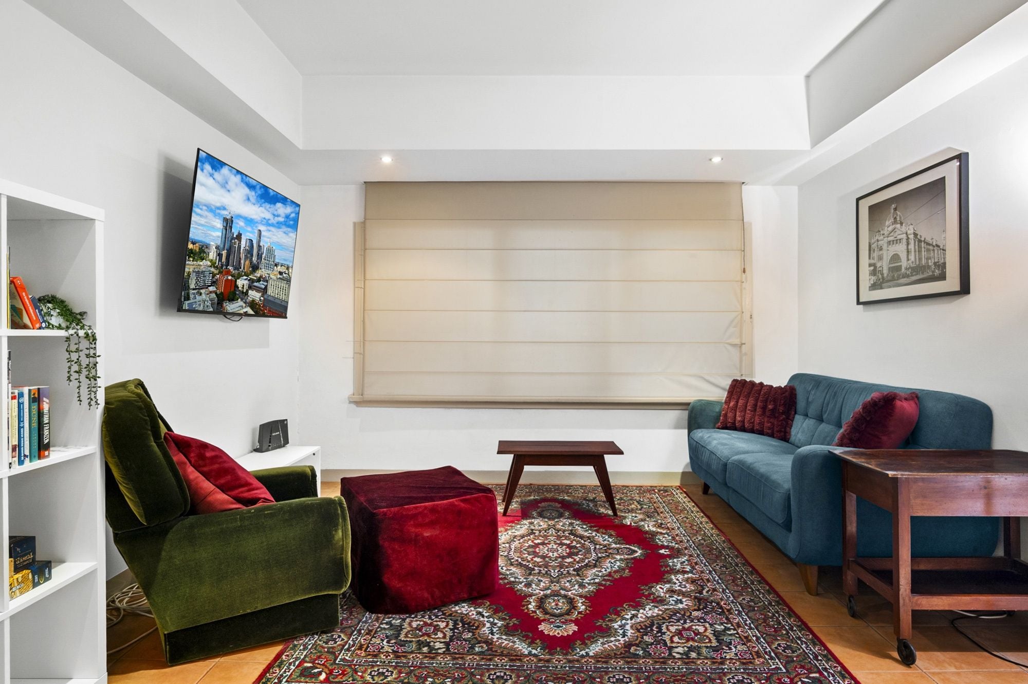 A modern living room with a sofa, armchair, and ottoman, a coffee table, a bookshelf, a large window with blinds, and a cityscape painting.