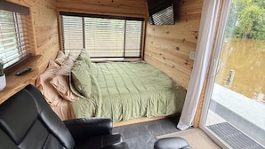 A wooden cabin interior with a bed, two black leather chairs, a TV mounted on the wall, and a window with blinds.