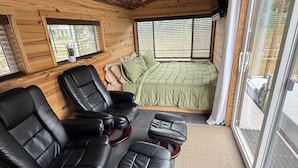 A cozy room with a wooden ceiling, a bed with green bedding, a seating area with leather chairs, and a large window.