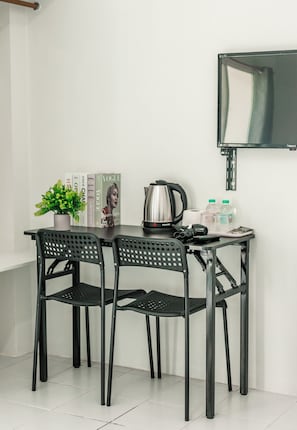 A small, modern kitchenette with a table, two chairs, a kettle, a camera, and a flat-screen TV mounted on the wall.