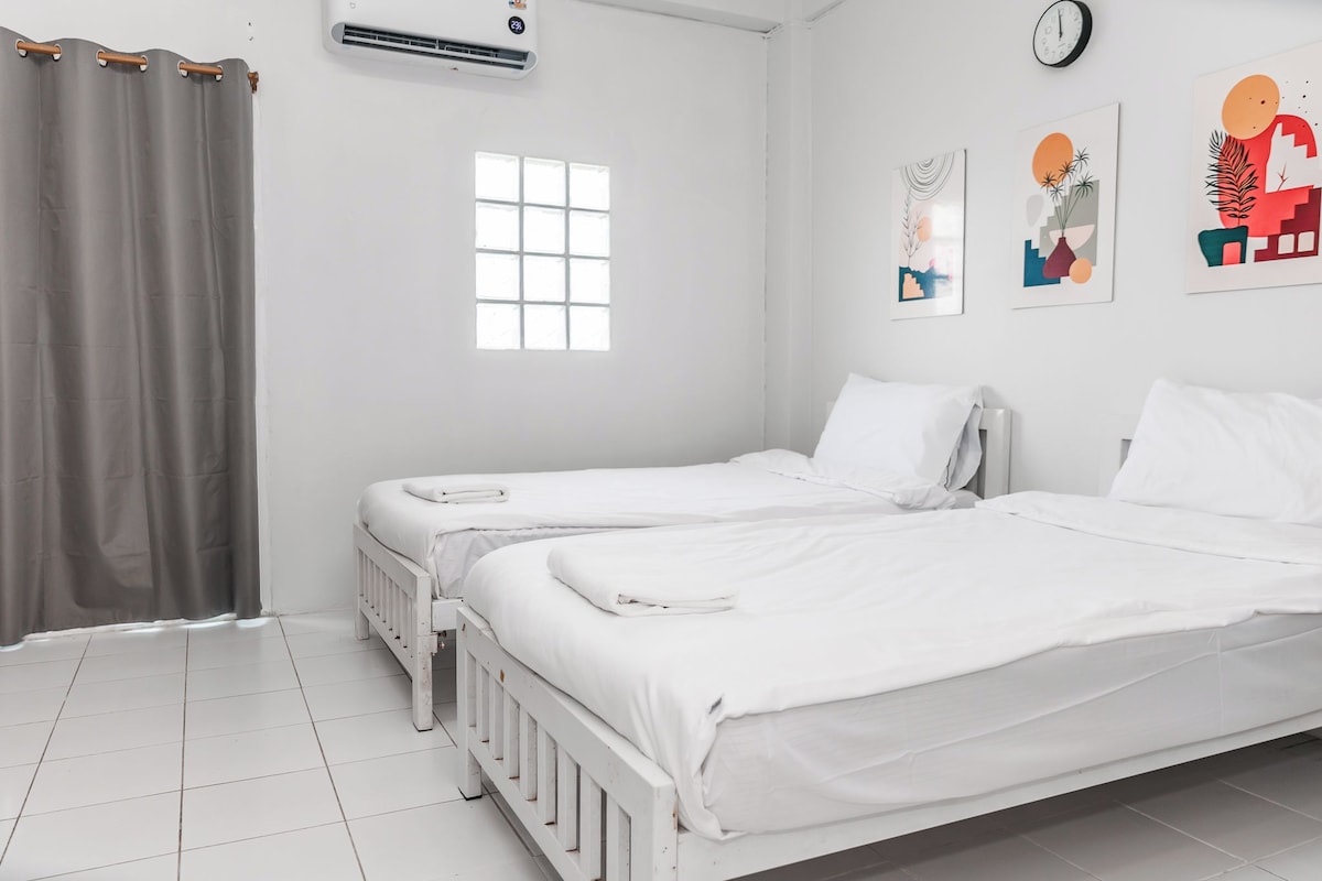 A hotel room with two single beds, a window, a clock, and a wall-mounted air conditioner.