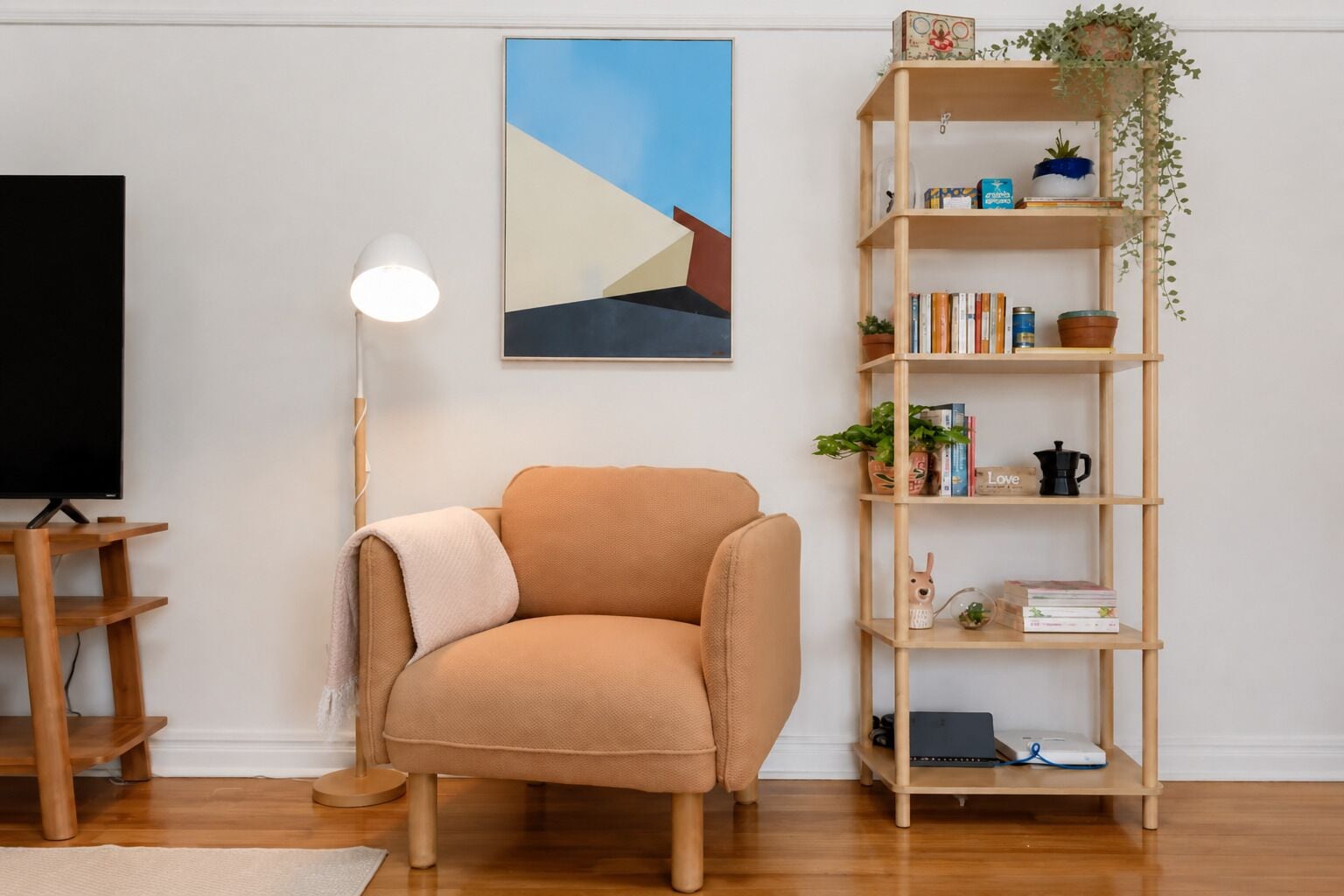 A living room with a tan sofa, a wooden bookshelf, a floor lamp, and a painting on the wall.