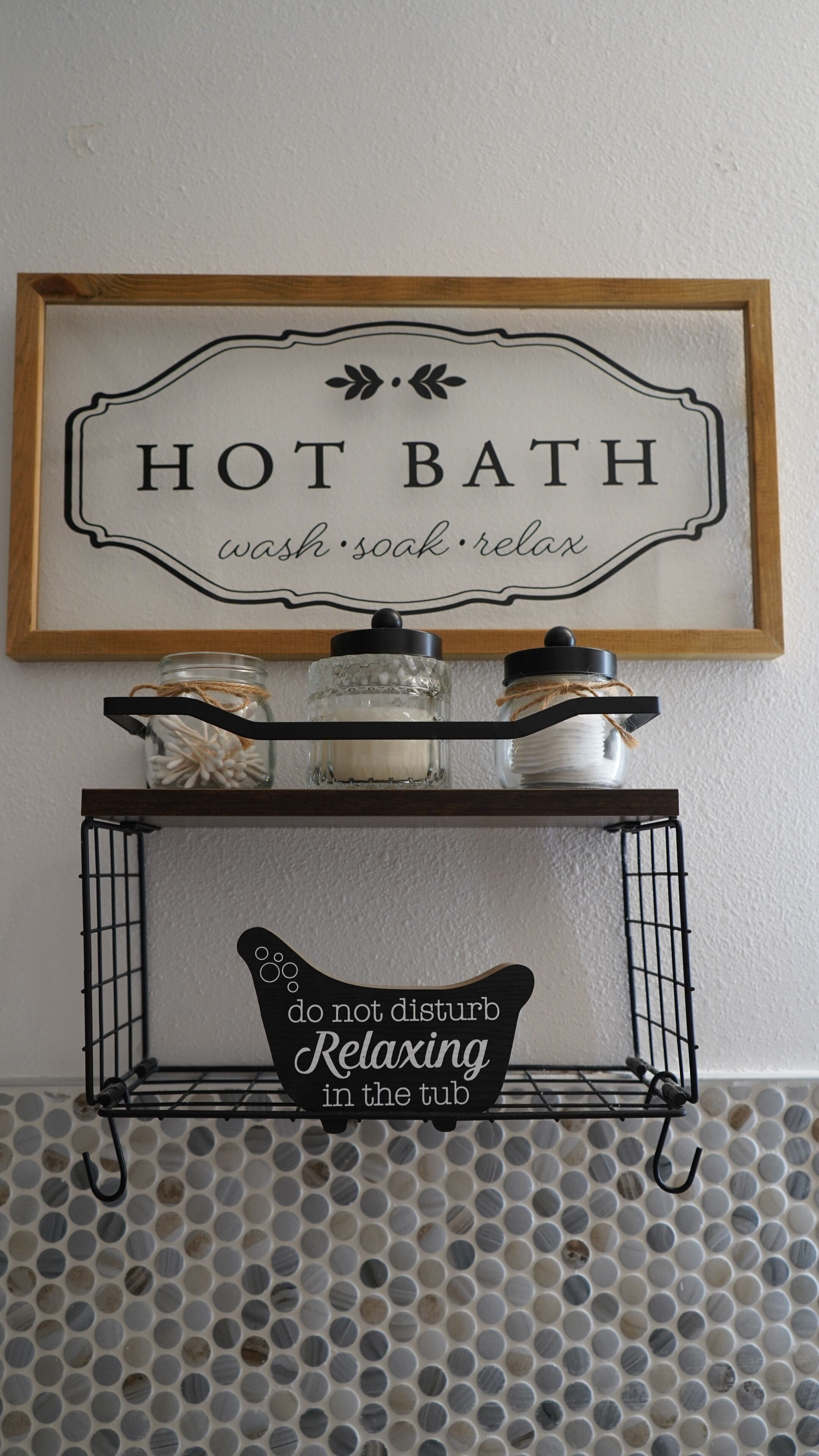 Ein gerahmtes Schild mit den Aufschriften 'HEISSES BAD' und 'waschen, baden, entspannen' über einem Regal mit Gläsern und einem Schild, das 'nicht stören, entspanne ich im Bad' sagt.