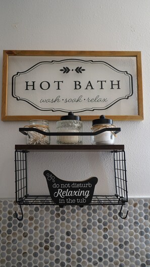 A framed sign with the words 'HOT BATH' and 'wash soak relax' above a shelf with jars and a sign that says 'do not disturb relaxing in the tub'.