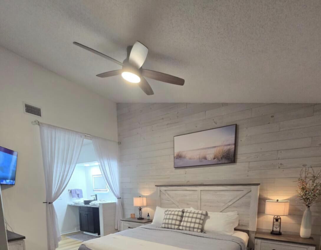 Ein Schlafzimmer mit einem Bett, Deckenventilator, Nachttisch mit Lampe, Gemälde an der Wand und Fenster mit Vorhängen.