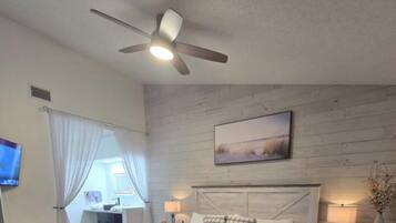 Ein Schlafzimmer mit einem Bett, Deckenventilator, Nachttisch mit Lampe, Gemälde an der Wand und Fenster mit Vorhängen.