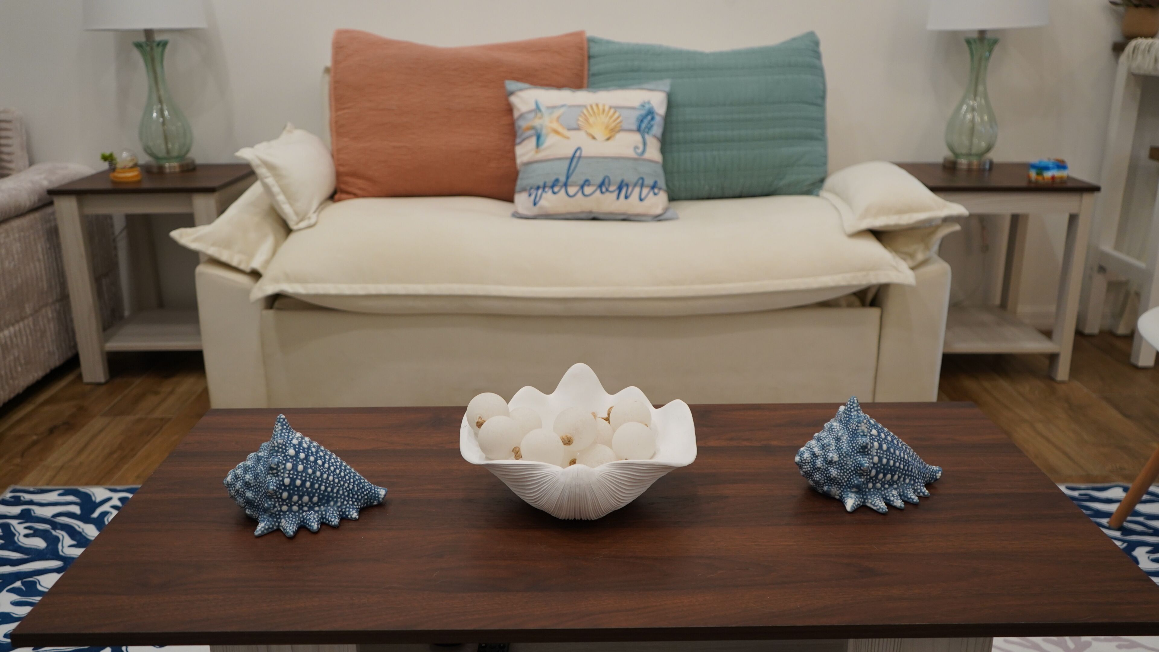 Ein Wohnzimmer mit einem beigen Sofa, einem 'welcome'-Kissen und dekorativen Muscheln auf einem Holztisch.