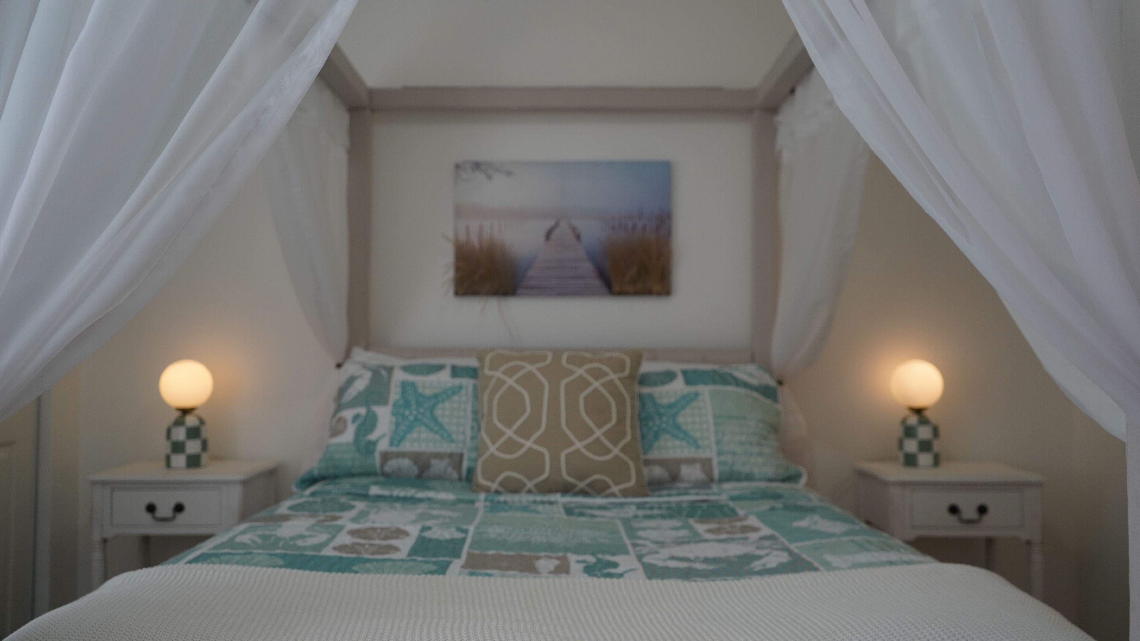 A bedroom with a bed, two bedside tables, a nightstand, a painting on the wall, and a canopy over the bed.