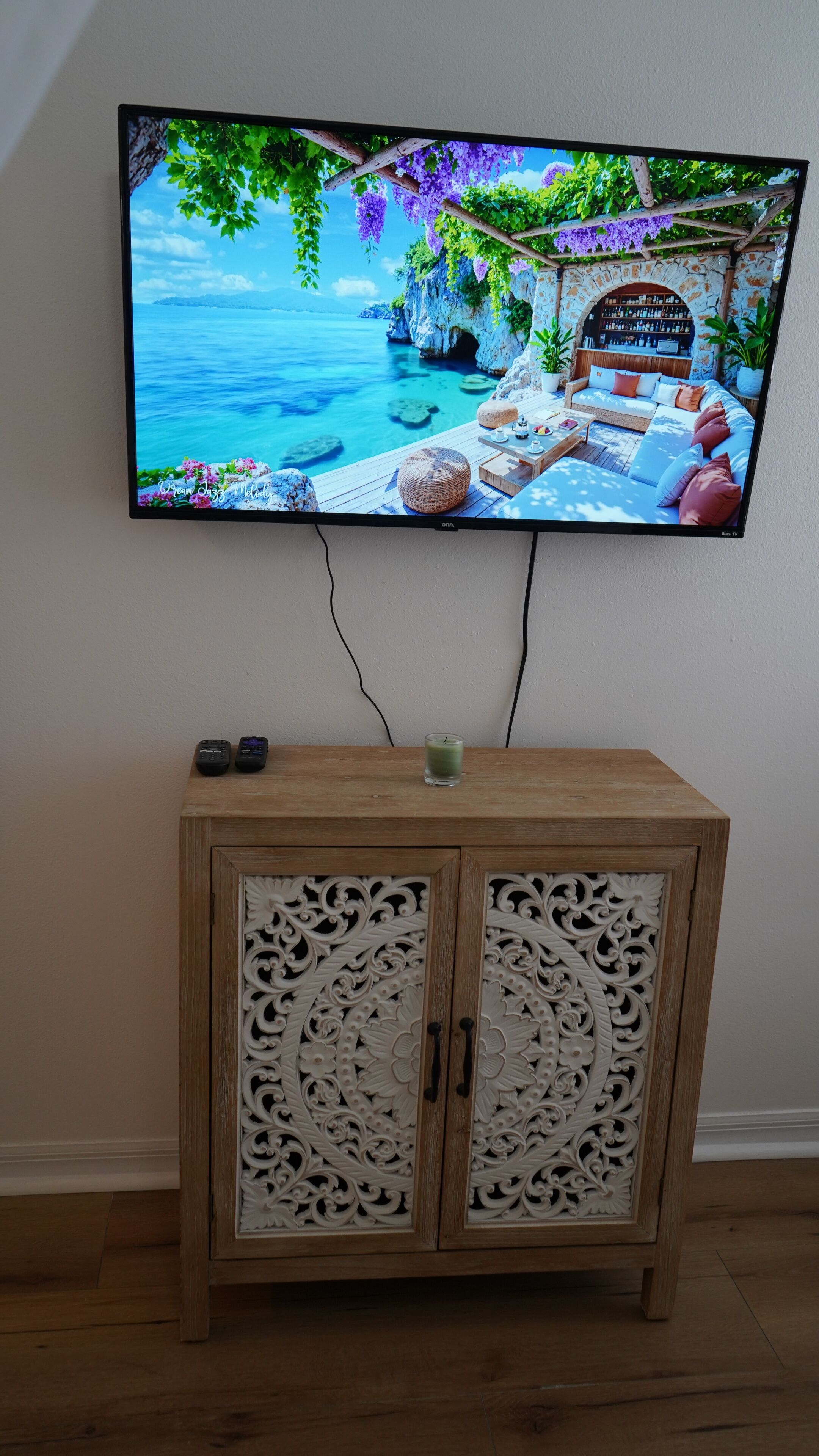 A television mounted on a wall displaying a scenic view of a coastal area with a wooden cabinet below.