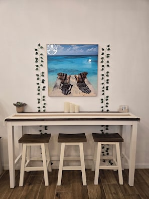 A wall-mounted painting of a beach scene with two Adirondack chairs, a small potted plant, and a candle holder on a white table with three wooden stools.