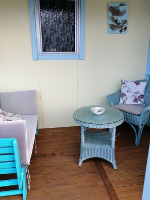 A room with a round glass table, a blue wicker chair, a sofa, and a window with a painting. - Relax, Coastal and Warm-Top Deck Beach House (Pataua South)