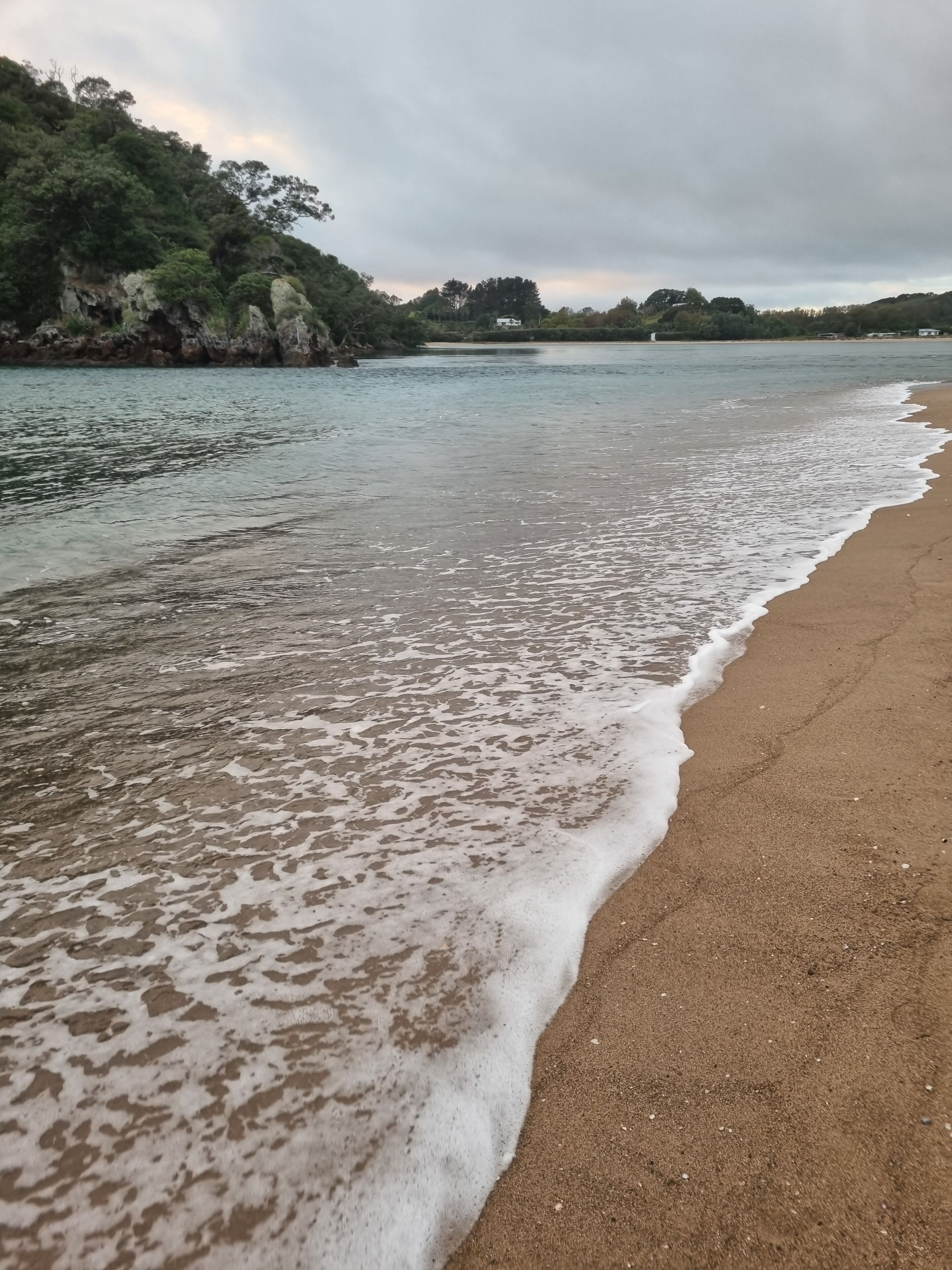 A beach with waves gently lapping onto the shore, a clear sky, and a hilly landscape in the background.