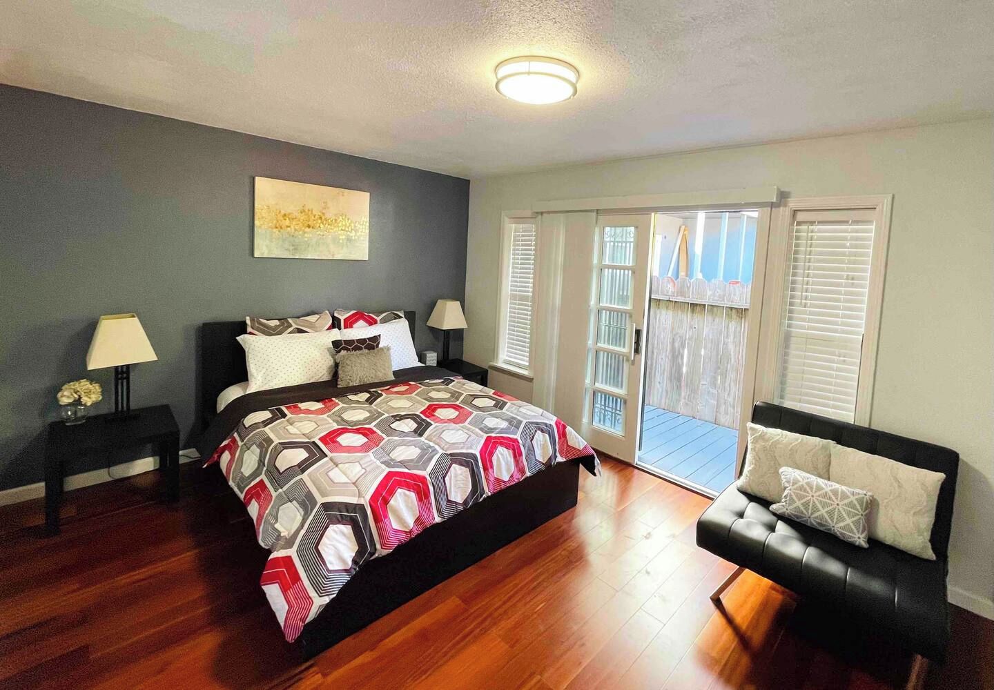 A bedroom with a large bed, a nightstand, a chair, a sliding glass door to a balcony, and a ceiling light.