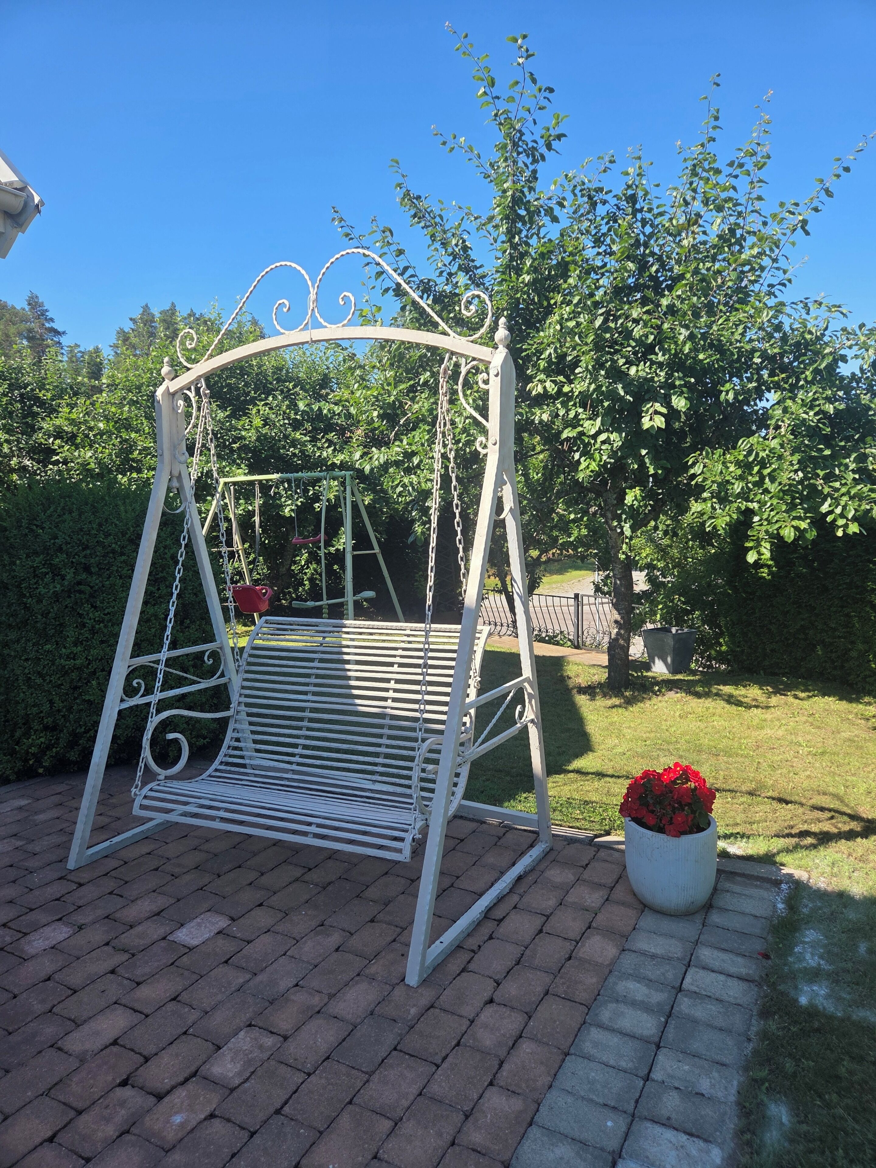 A metal swing set with a bench, surrounded by a brick pathway and greenery.
