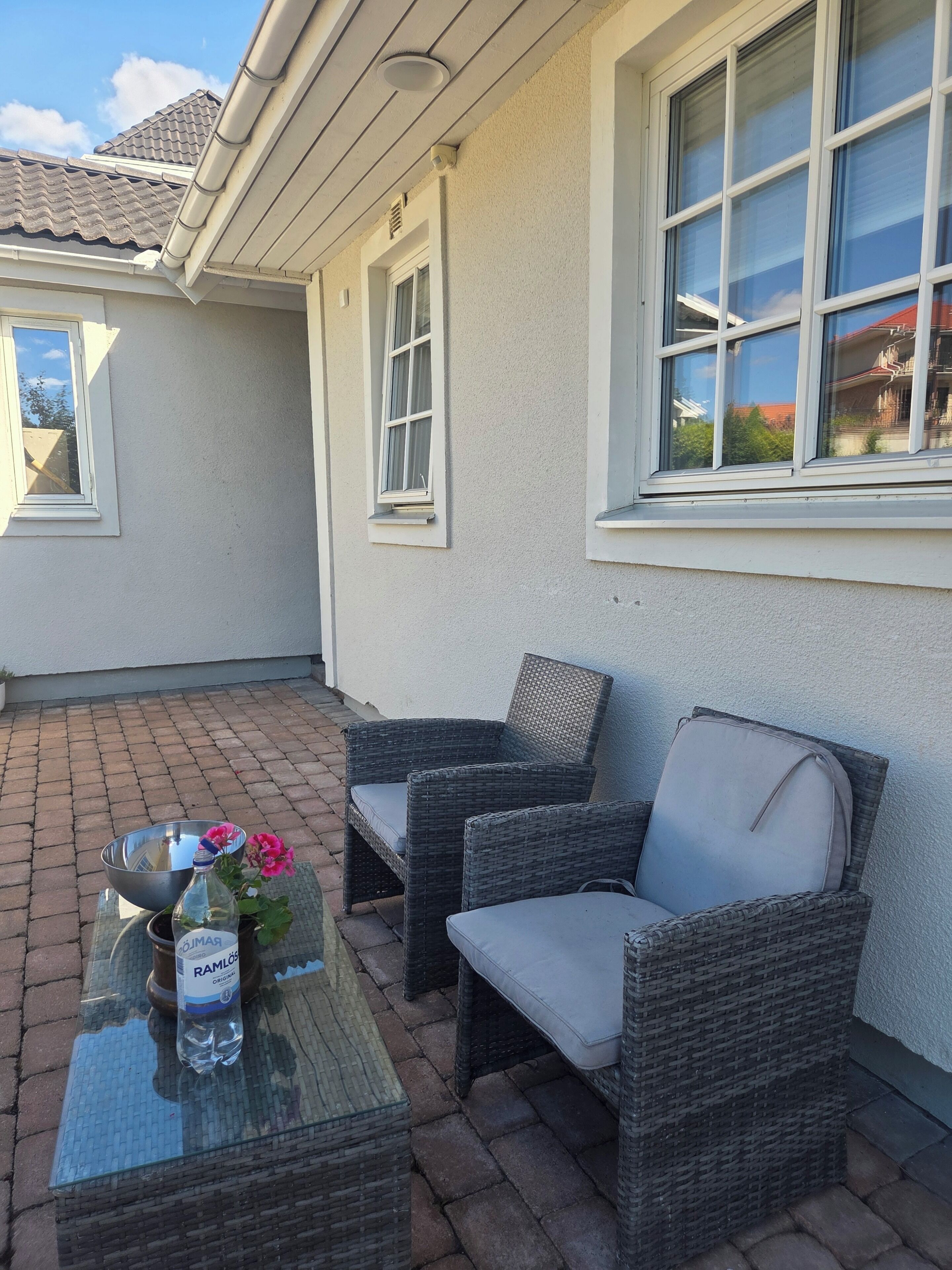 A patio area with wicker furniture, a glass table, and a metal bowl with flowers.