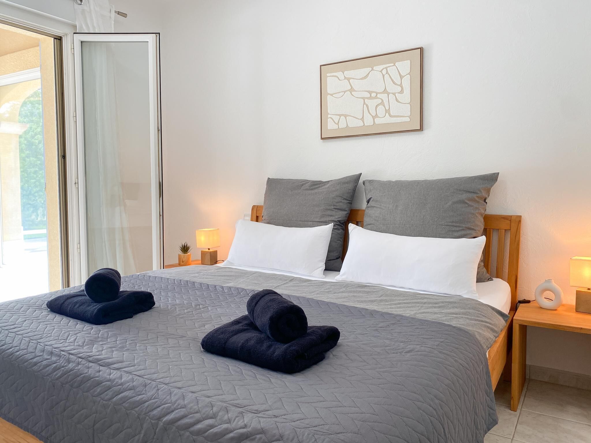 Ett hotellrum med en säng, kuddar, ett sängbord med en lampa och ett konstverk som hänger på väggen.