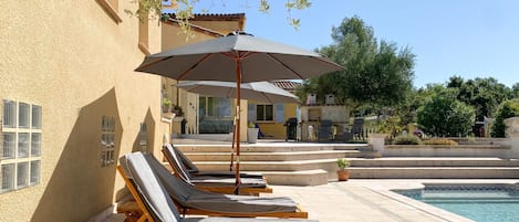 A poolside area with lounge chairs and umbrellas, a pool, and a building with a tiled roof.