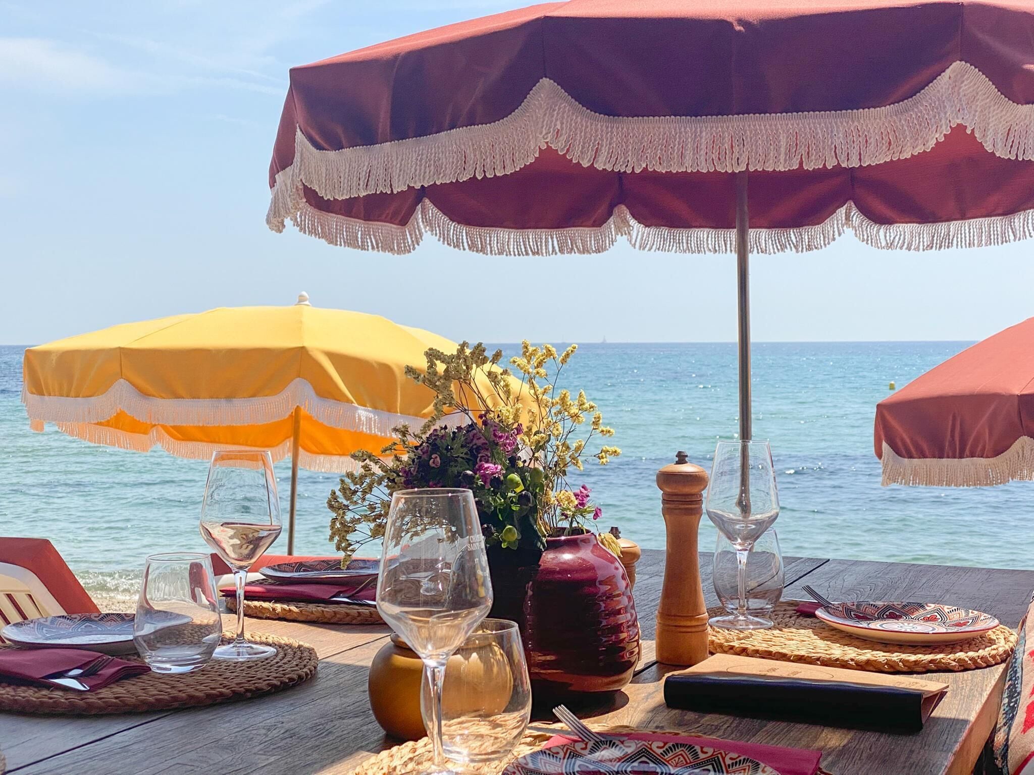 A table set for a meal with wine glasses, a salt shaker, and a vase of flowers, under beach umbrellas with a view of the sea.