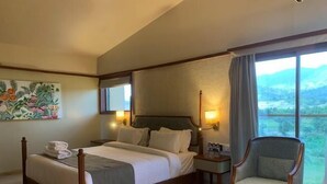 A hotel room with a bed, a chair, a small table, a painting on the wall, and a window with a view. - Vinti Resort, Ambaji (Danta)