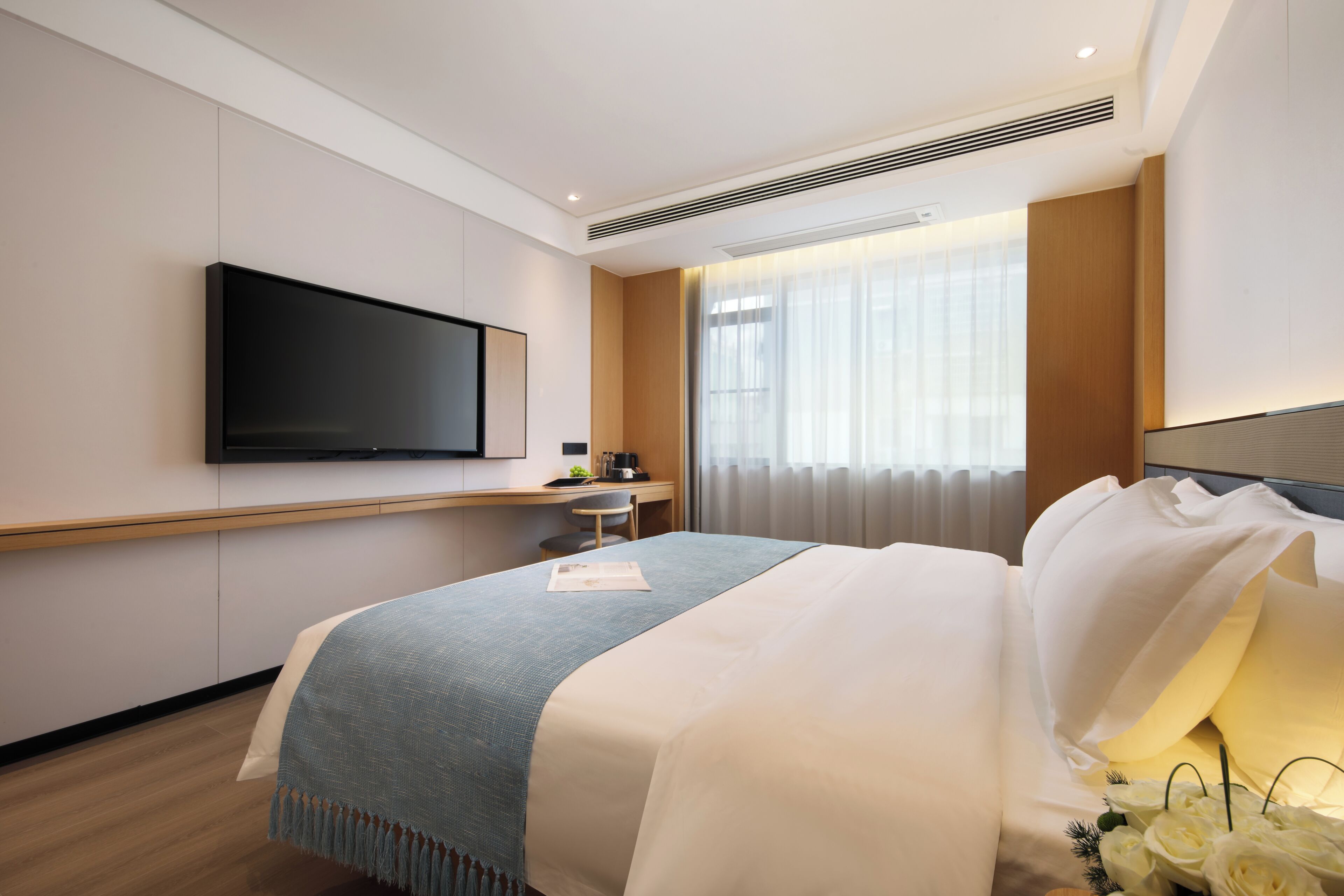 Una habitación de hotel con una cama, un televisor de pantalla plana montado en la pared, una mesita con una cafetera y una ventana con cortinas.