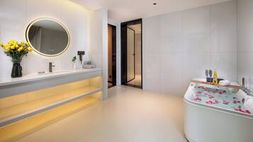 A modern bathroom with a vanity, a large mirror, a bathtub with rose petals, and a door leading to another room.